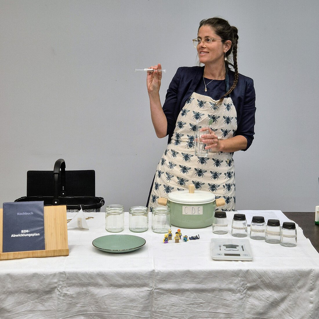 Eine Frau steht hinter einem Tisch, auf dem verschiedene Gläser und Behälter mit verschiedenen Inhalten stehen. Sie erklärt etwas und gestikuliert dabei. Sie lächelt.