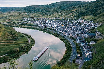 Ein Weinort an der Mosel, mit einem Lastkahn, der den Fluss entlangfährt. Die sanfte Landschaft ist von Weinbergen und Hügeln umgeben.