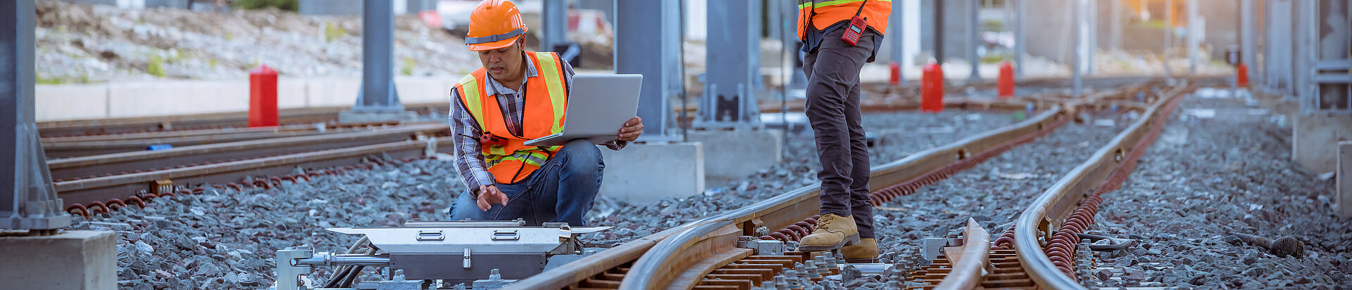 Zwei Bauarbeiter stehen an einer Bahnanlage. Eine Person hockt am Gleisen und die andere steht zwischen Gleisen und überprüft die Anlage mit Hilfe von BIM.