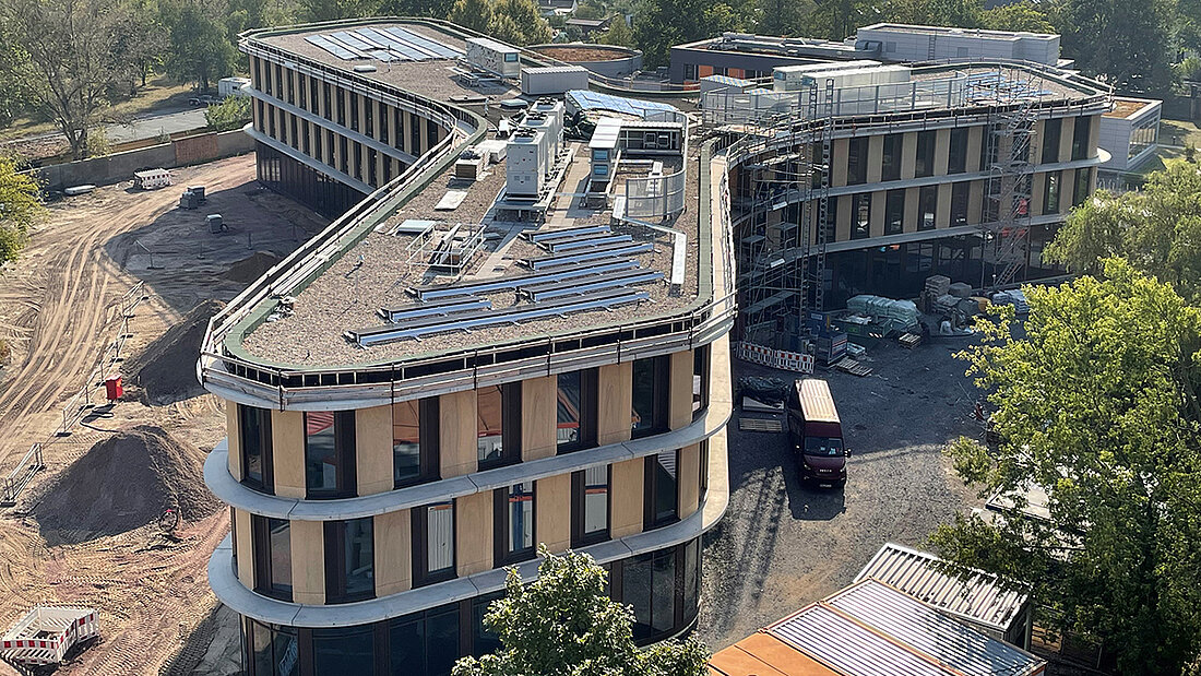 Das Bild zeigt eine moderne Gebäudestruktur mit geschwungener Form in der Bauphase. Auf dem Dach sind technische Anlagen und Montagestrukturen zu sehen, vermutlich für eine Photovoltaikanlage. Das Gebäude ist teilweise eingerüstet, Baumaterialien lagern im Hof, die Umgebung ist grün und städtisch geprägt.