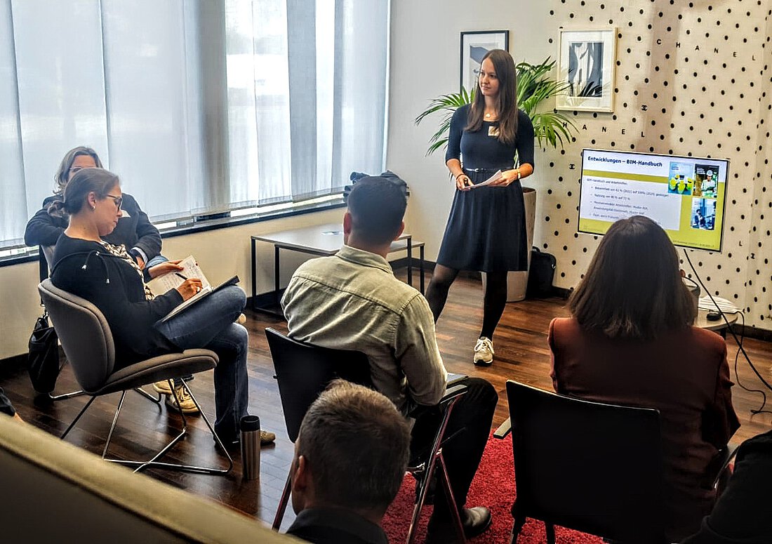 Eine Frau steht neben einem Bildschirm und erklärt etwas.Vor ihr sitzen mehrere Menschen im Publikum und hören konzentriert zu.