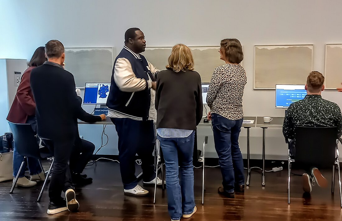 Es ist ein Raum mit verschiedenen Stationen zu sehen, an denen Computer stehen. Menschen stehen an den Stationen und unterhalten sich angeregt. 